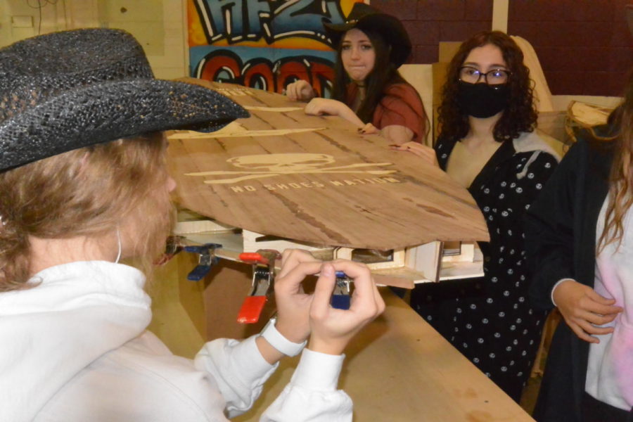 While junior Abigail Bartold and freshman Kaelyn Tracy hold a strip in place, senior Heather Gravely glues and clamps the rails on a paddleboard the Henry Ford II students are creating for country music star Kenny Chesney.